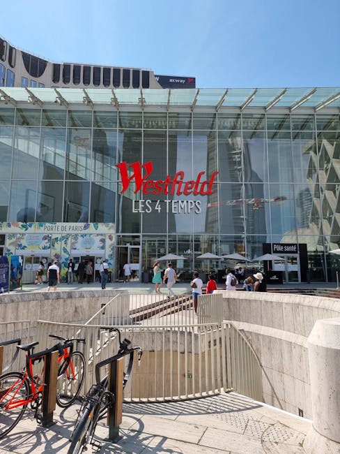 The exterior of Westfield shopping centre at Shepherds Bush, featuring large glass façade and prominent red Westfield sign, with people walking on the pavement and entering the building. In the foregr